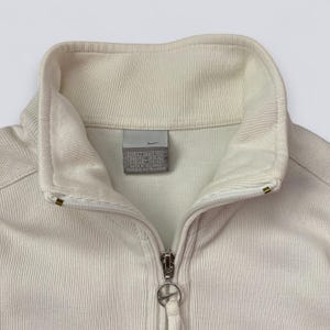 以下が含まれることがあります： クリーム色のナイキジップアップジャケット、ハイカラー。 ジャケットは、ナイキのロゴプル付きのシルバーのジッパーと小さなナイキのロゴタグが特徴です。 生地はリブ編みのテクスチャです。