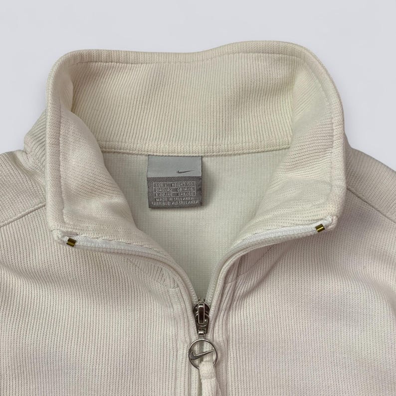 以下が含まれることがあります： クリーム色のナイキジップアップジャケット、ハイカラー。 ジャケットは、ナイキのロゴプル付きのシルバーのジッパーと小さなナイキのロゴタグが特徴です。 生地はリブ編みのテクスチャです。