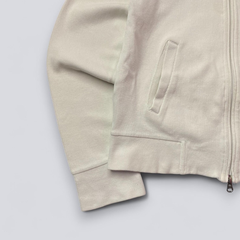 以下が含まれることがあります： リブ編みのテクスチャが特徴の、ライトベージュのジップアップスウェットシャツ。スウェットシャツにはフロントポケットとフルレングスのジッパーが付いています。袖口と裾はリブ編みです。