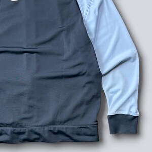 May include: A dark gray and light blue athletic jacket. The jacket has a dark gray body and light blue sleeves. The cuffs and bottom hem are dark gray. The jacket appears to be made of a smooth, athletic material.