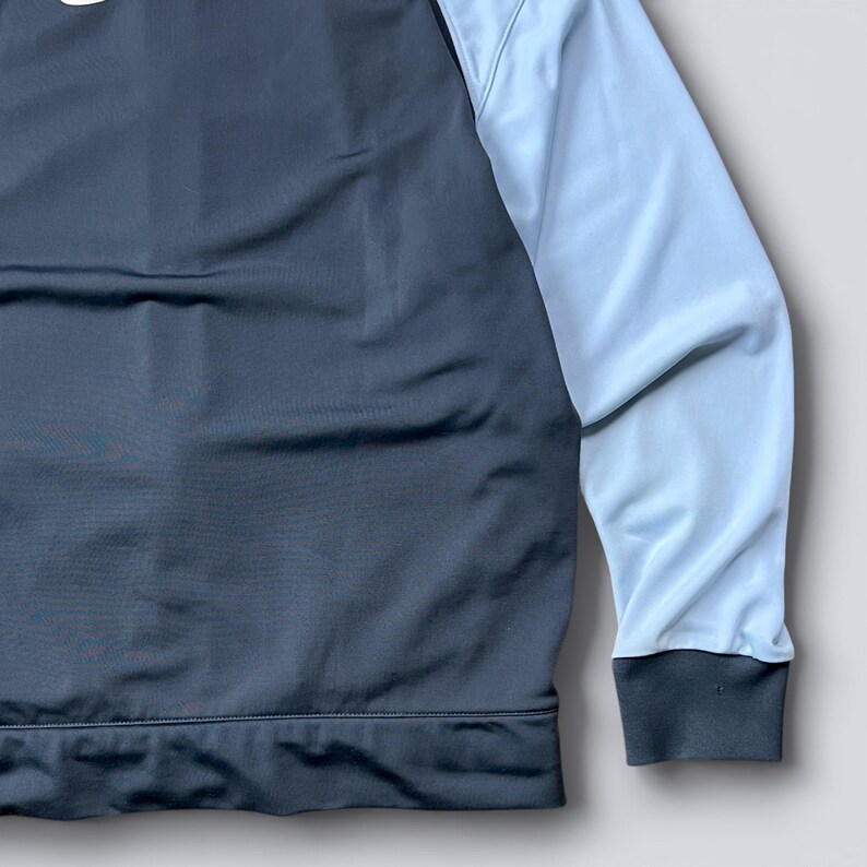 May include: A dark gray and light blue athletic jacket. The jacket has a dark gray body and light blue sleeves. The cuffs and bottom hem are dark gray. The jacket appears to be made of a smooth, athletic material.
