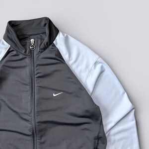 May include: Black and light blue track jacket with a full zipper and a stand-up collar. The jacket features a white Nike swoosh logo on the chest. The sleeves are light blue, and the body is black.