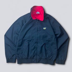 May include: Navy blue zip-up jacket with a red collar lining. The jacket features a CAT logo on the chest and two front pockets. The jacket has a ribbed hem and cuffs.