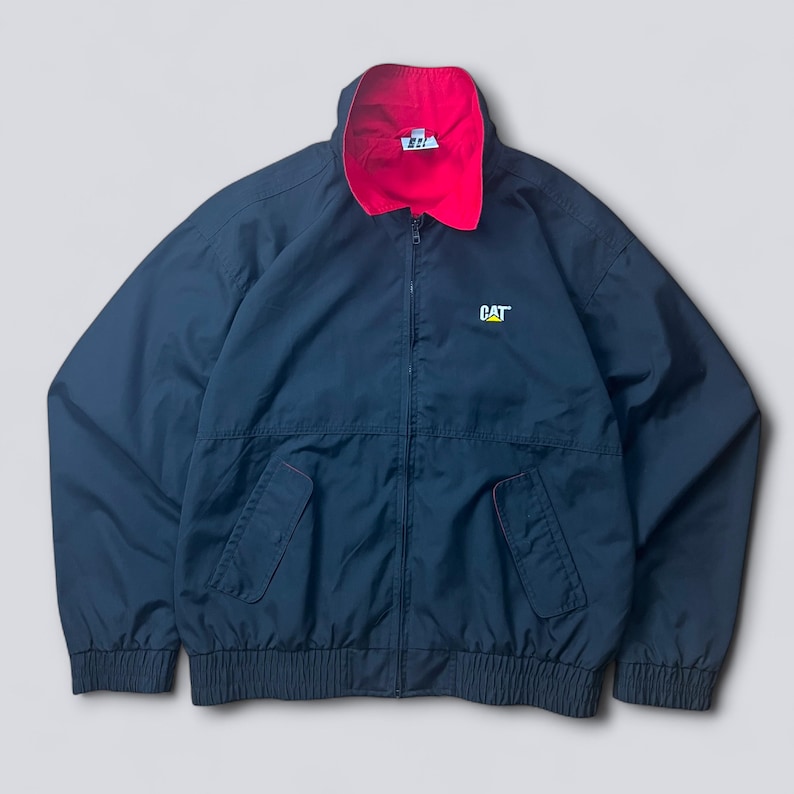 May include: Navy blue zip-up jacket with a red collar lining. The jacket features a CAT logo on the chest and two front pockets. The jacket has a ribbed hem and cuffs.