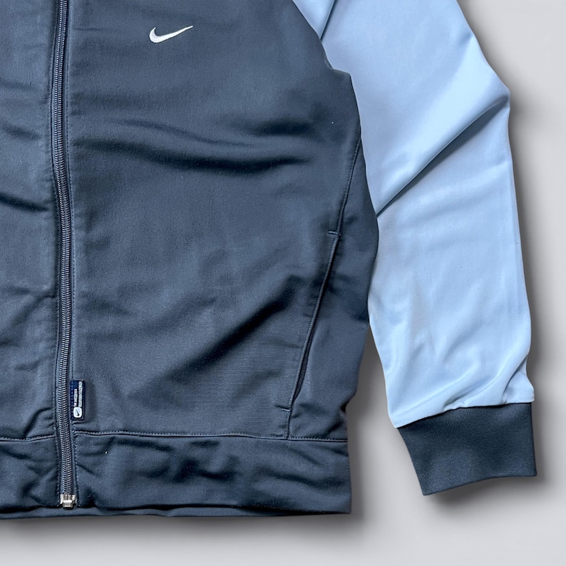 May include: A dark gray and light blue track jacket with a full zipper closure. The jacket features a small white Nike logo on the chest and a light blue sleeve. The jacket has a dark gray cuff and a pocket.