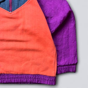 May include: A color-blocked pullover jacket featuring a bright orange fleece front panel, deep purple crinkled nylon sleeves, and a matching purple elasticized hem. The upper chest area is dark blue with a central vertical zipper closure.