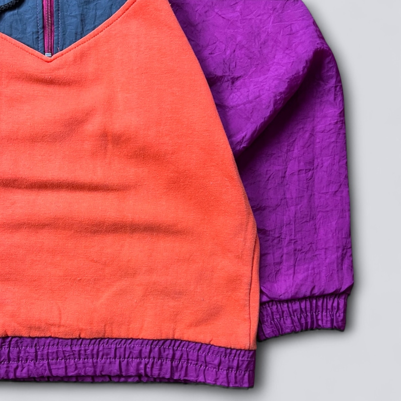 May include: A color-blocked pullover jacket featuring a bright orange fleece front panel, deep purple crinkled nylon sleeves, and a matching purple elasticized hem. The upper chest area is dark blue with a central vertical zipper closure.