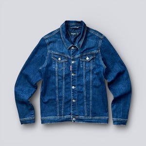 May include: A classic blue denim jacket with a button-up front, two chest pockets, and side pockets. The jacket has a collared neckline and long sleeves. A small red tag is visible on the left chest pocket. The jacket is made of a sturdy denim material.