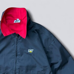 May include: Black zip-up jacket with a red collar lining. The jacket features a CAT logo on the front and a small logo on the collar. The jacket has a full zipper and a hood.