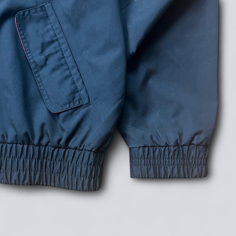 May include: Close-up of a navy blue jacket with a red-lined pocket. The jacket has elastic cuffs and a ribbed hem. The fabric appears to be a smooth, possibly water-resistant material.