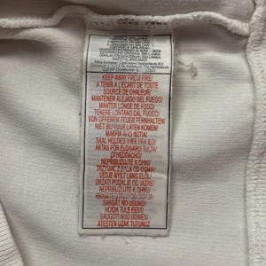 以下が含まれることがあります： 黒い文字が入った白い布タグのクローズアップ。タグには、複数の言語で「KEEP AWAY FROM FIRE」というテキストが含まれています。タグにはナイキのロゴと製造情報も含まれています。