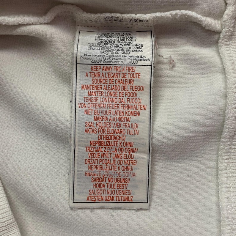 以下が含まれることがあります： 黒い文字が入った白い布タグのクローズアップ。タグには、複数の言語で「KEEP AWAY FROM FIRE」というテキストが含まれています。タグにはナイキのロゴと製造情報も含まれています。