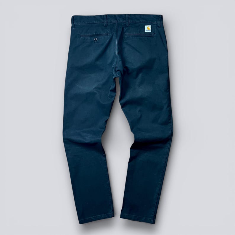 May include: Navy blue pants with a classic fit. The pants feature a button closure, belt loops, and back pockets. A small, square logo is visible on the back pocket. The pants are made of a durable fabric.