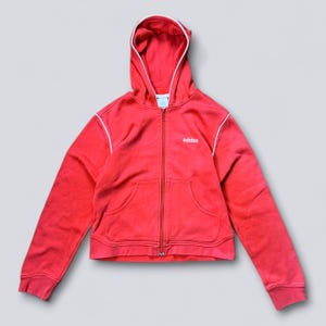 Peut inclure: Sweat à capuche rouge zippé avec capuche et deux poches avant. Le sweat à capuche a une bordure blanche le long de la capuche et de la fermeture éclair, et le logo Adidas est sur la poitrine. Le sweat à capuche est en matière douce.