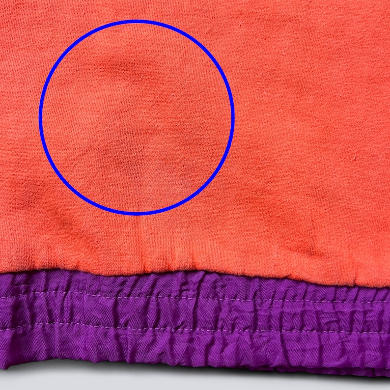 May include: A bright coral orange fabric section featuring a prominent blue circular outline highlighting a faint, darker discoloration on the material. Below the coral section is a contrasting, textured purple hem with stitching.