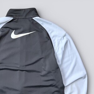 May include: A dark gray and light blue athletic jacket with a white Nike logo. The jacket has a high collar and white sleeves. The Nike swoosh is prominently displayed on the back of the jacket.