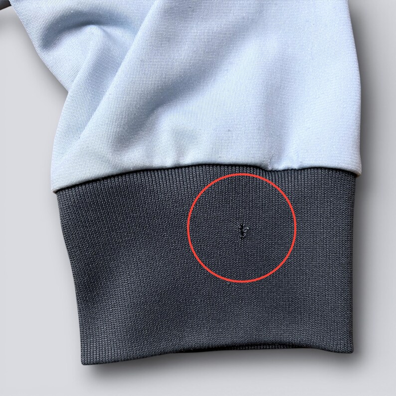 May include: Close-up of a light blue sweatshirt sleeve with a dark gray ribbed cuff. A small hole is visible on the cuff. The fabric appears soft and slightly wrinkled.
