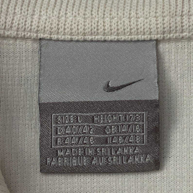 以下が含まれることがあります： 白の衣服に縫い付けられた灰色のナイキタグのクローズアップ。タグにはナイキのスウッシュロゴと、サイズ、高さ、測定値を含むテキストがあります。タグには「Made in Sri Lanka」と「Fabriqu&eacute; au Sri Lanka」と書かれています。