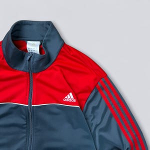 May include: A red and navy blue Adidas track jacket with a white Adidas logo on the chest. The jacket has red stripes down the sleeves and a white stripe along the zipper.
