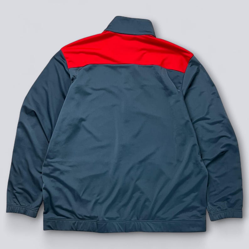 May include: A teal and red track jacket. The jacket has a red panel across the shoulders and upper back, with a teal body and sleeves. It features a stand-up collar and a full-length zipper.