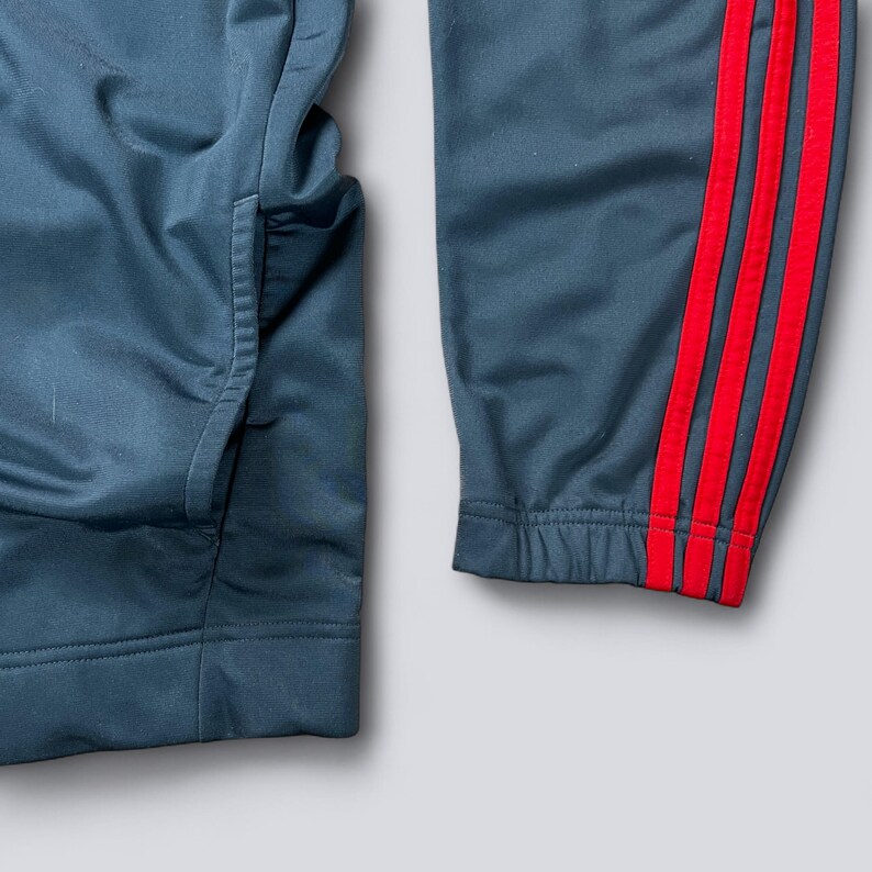 May include: Close-up of dark teal athletic pants with a red stripe design on the side. The pants have a pocket on the left side and a ribbed cuff at the bottom. The fabric has a slight sheen.