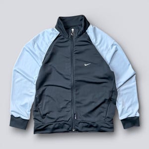 May include: A black and light blue track jacket with a full zipper closure and a small white Nike logo on the chest. The jacket has light blue sleeves and a black body, with black trim on the cuffs and collar.