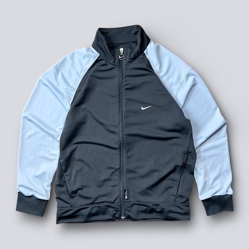 May include: A black and light blue track jacket with a full zipper closure and a small white Nike logo on the chest. The jacket has light blue sleeves and a black body, with black trim on the cuffs and collar.