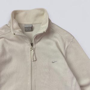 以下が含まれることがあります： クリーム色のジップアップジャケット、ハイカラー。 ジャケットはシルバーのジッパーと胸に小さなナイキのロゴが付いています。 生地はリブ編みのテクスチャです。
