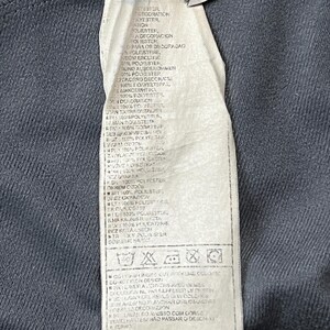 May include: Close-up of a clothing tag with text in multiple languages. The tag is white with black text and is attached to a dark gray fabric. The tag includes care instructions and fabric composition details.