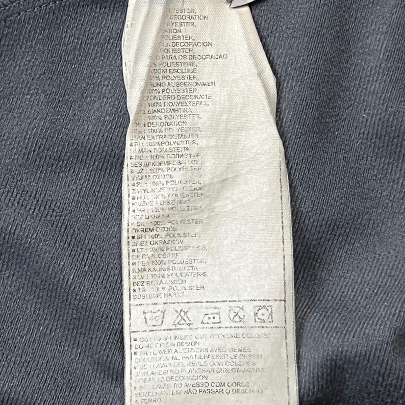 May include: Close-up of a clothing tag with text in multiple languages. The tag is white with black text and is attached to a dark gray fabric. The tag includes care instructions and fabric composition details.