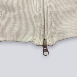 以下が含まれることがあります： 白いニットセーターのクローズアップ。銀色のジッパーが付いています。ジッパーは中央に配置され、完全に閉じられており、金属製のプルタブが見えています。ニット生地はリブ編みのテクスチャです。