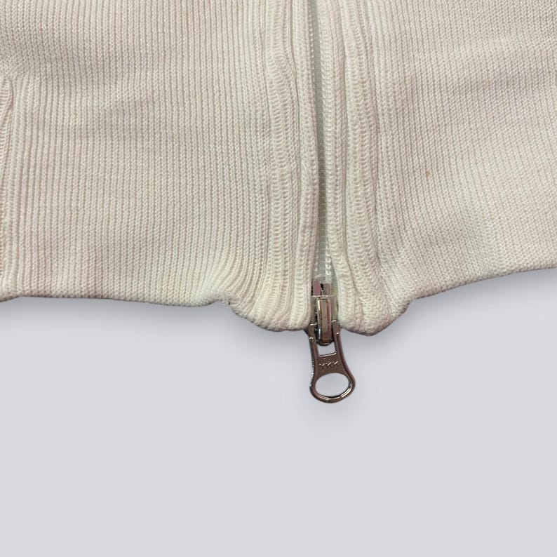 以下が含まれることがあります： 白いニットセーターのクローズアップ。銀色のジッパーが付いています。ジッパーは中央に配置され、完全に閉じられており、金属製のプルタブが見えています。ニット生地はリブ編みのテクスチャです。