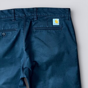 May include: Close-up of navy blue pants with a small square Carhartt logo patch. The pants have two back pockets and a button closure. The fabric appears to be a sturdy cotton blend, suitable for work or casual wear.