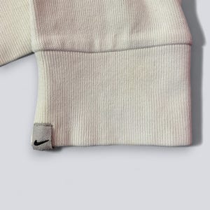 以下が含まれることがあります： クリーム色のスウェットシャツの袖口のクローズアップ。リブ編みのテクスチャです。黒いナイキのスウッシュロゴが付いた小さな灰色のタグが袖口に付いています。袖口は折り返されており、内側の生地が見えています。