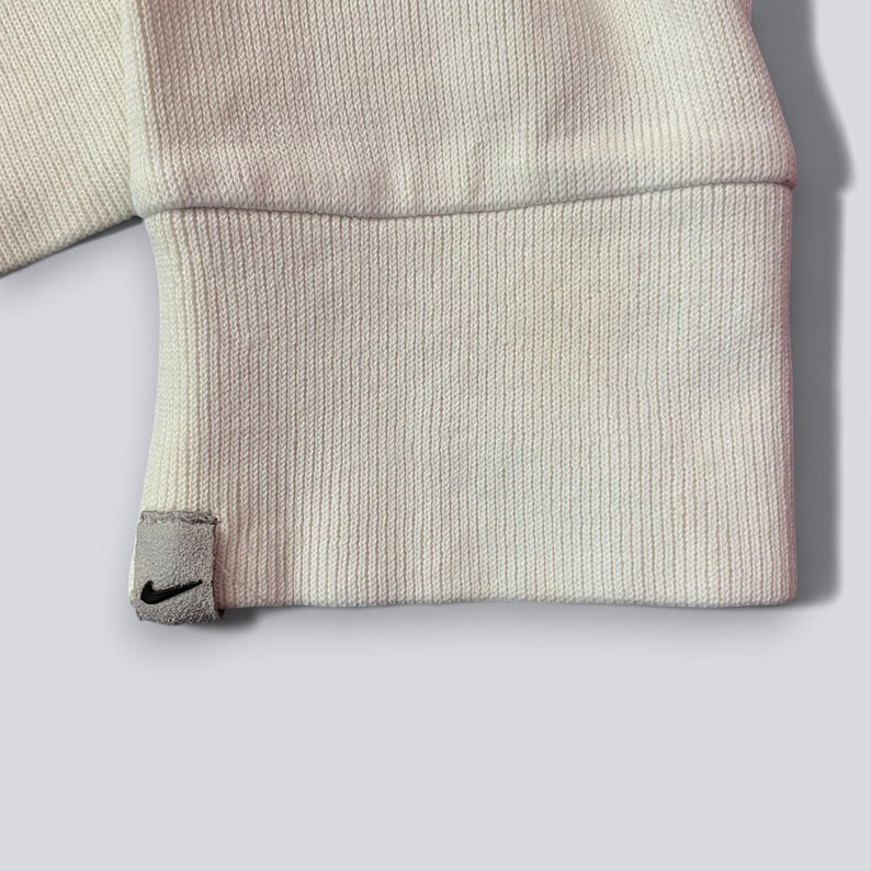 以下が含まれることがあります： クリーム色のスウェットシャツの袖口のクローズアップ。リブ編みのテクスチャです。黒いナイキのスウッシュロゴが付いた小さな灰色のタグが袖口に付いています。袖口は折り返されており、内側の生地が見えています。