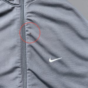 May include: Close-up of a gray Nike track jacket with a full-length zipper. The jacket features a white Nike swoosh logo on the chest. The fabric has a subtle sheen. There is a small stain near the zipper.