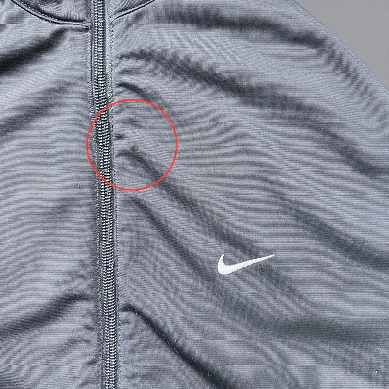 May include: Close-up of a gray Nike track jacket with a full-length zipper. The jacket features a white Nike swoosh logo on the chest. The fabric has a subtle sheen. There is a small stain near the zipper.
