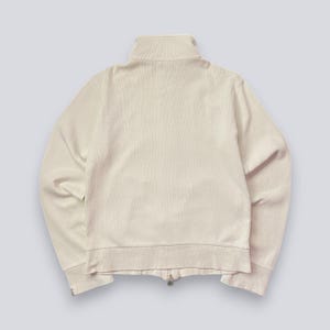 以下が含まれることがあります： クリーム色のジップアップジャケット、ハイカラー付き。 ジャケットはリブ編みのテクスチャで、フルレングスのジッパーが付いています。 袖口と裾もリブ編みです。 明るい背景に対して、ジャケットの後ろ姿が写っています。