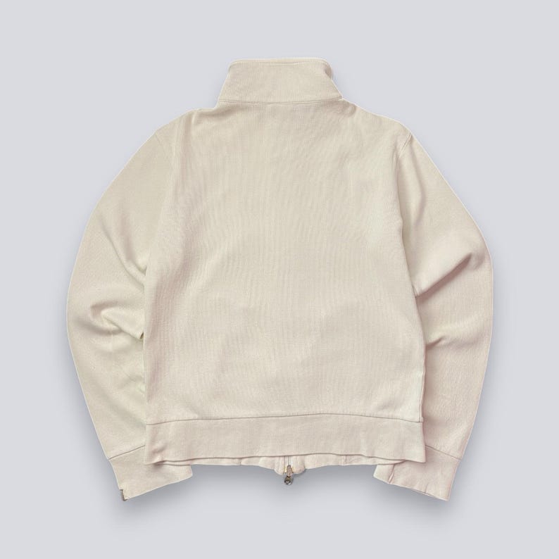 以下が含まれることがあります： クリーム色のジップアップジャケット、ハイカラー付き。 ジャケットはリブ編みのテクスチャで、フルレングスのジッパーが付いています。 袖口と裾もリブ編みです。 明るい背景に対して、ジャケットの後ろ姿が写っています。