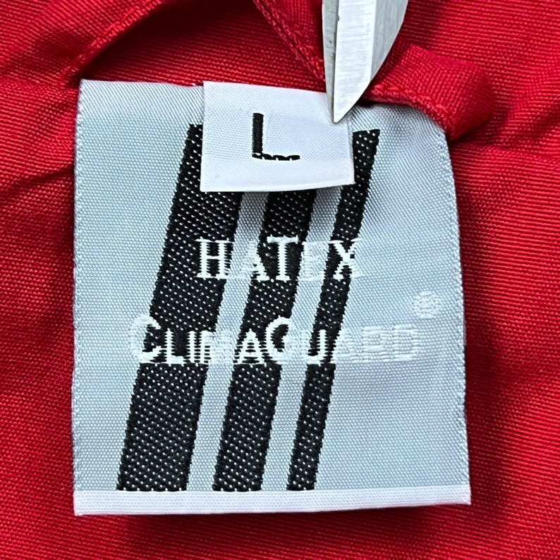 May include: Close-up of a clothing tag with the text "HATIX CLIMAGUARD" and the letter "L" on a white rectangle. The tag has three black vertical stripes and is set against a red fabric background.