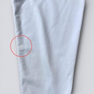 May include: Close-up of a light blue fabric, possibly clothing. The fabric has a subtle texture and a few small yellow spots. The image is well-lit, highlighting the details of the material.