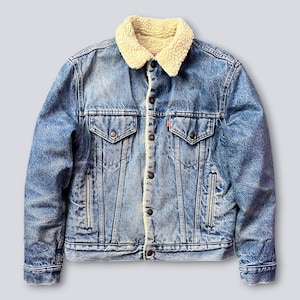 Può includere: Giacca di jeans blu con colletto e fodera in sherpa color crema. La giacca ha due tasche sul petto, due tasche laterali e una chiusura con bottoni. Una piccola etichetta rossa è visibile sulla tasca sul petto. Stile classico.