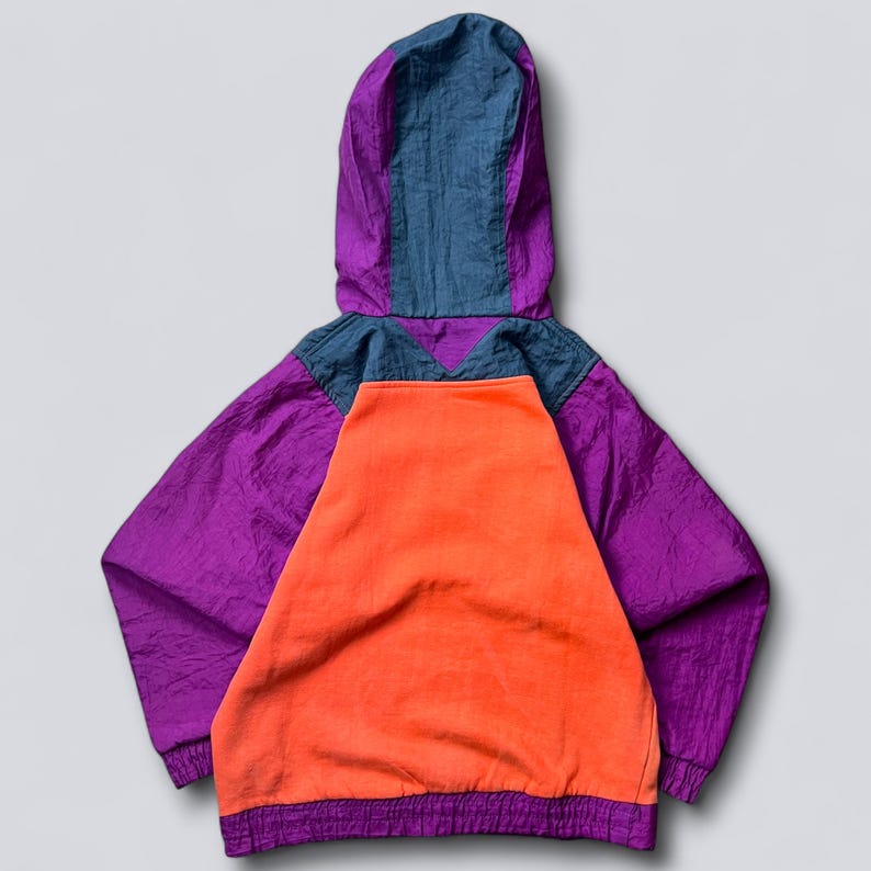 May include: A vintage hooded jacket in a vibrant color block design. The jacket features a purple body, an orange front panel, and a blue and purple hood. The jacket has a drawstring at the bottom and is made of a crinkled fabric.