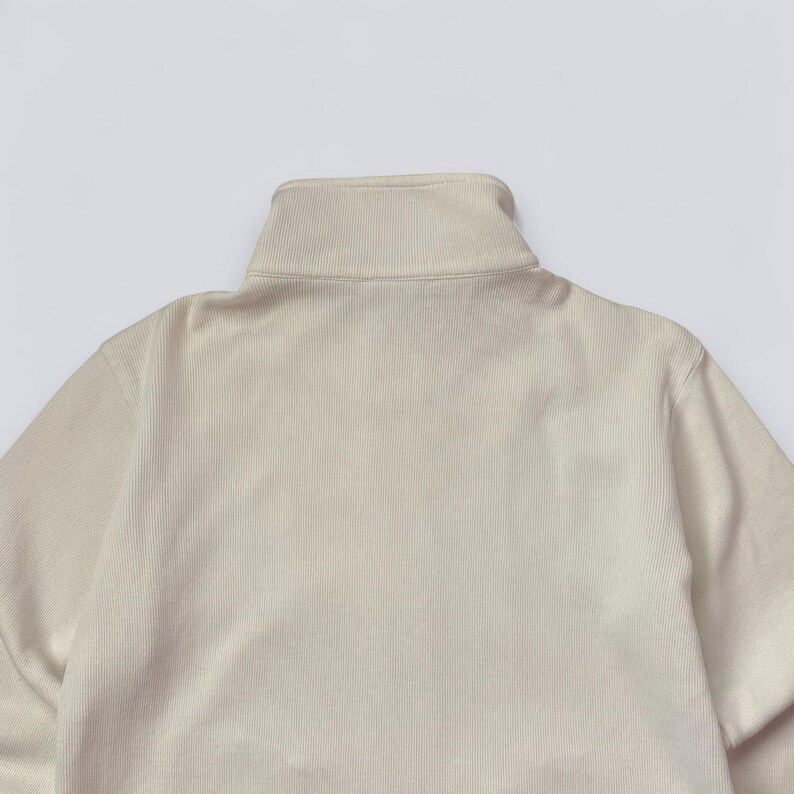 以下が含まれることがあります： ベージュのリブ編みプルオーバーセーターのクローズアップ。ハイネックの折り返し襟が特徴です。セーターはシンプルでミニマルなデザインで、生地は柔らかく快適に見えます。ニュートラルな色合いなので、さまざまなコーディネートに合わせやすいです。