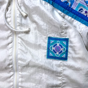 May include: A white vintage windbreaker jacket with a full zipper and drawstring hood. The jacket features blue and teal accents on the shoulders and a square patch with a nautical design. The jacket is made of a shiny, lightweight material.