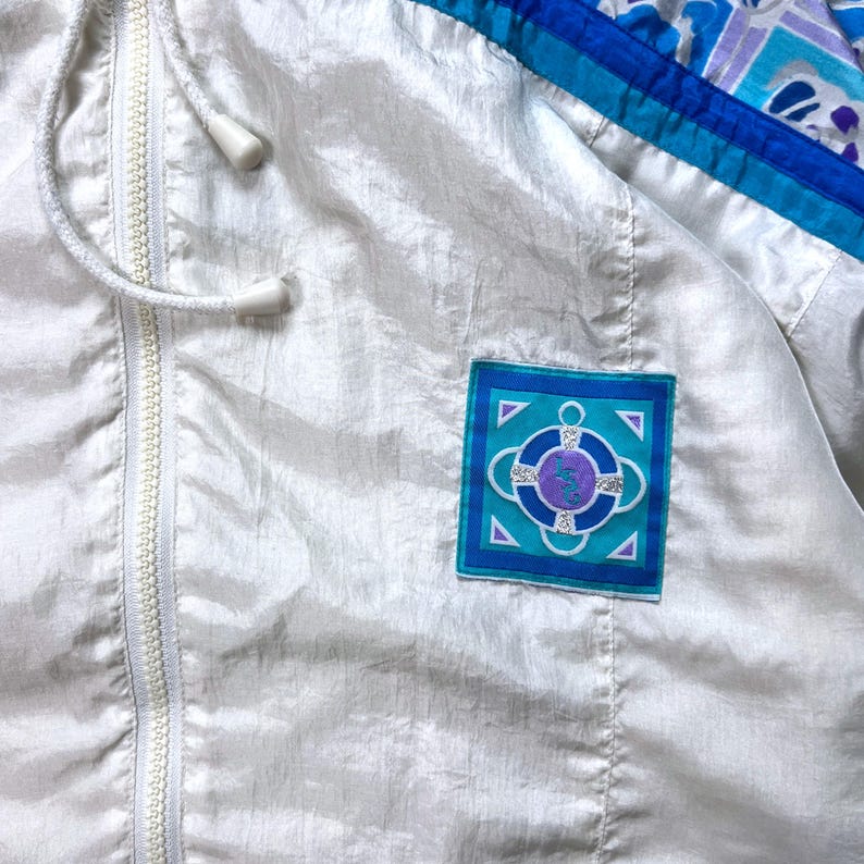 May include: A white vintage windbreaker jacket with a full zipper and drawstring hood. The jacket features blue and teal accents on the shoulders and a square patch with a nautical design. The jacket is made of a shiny, lightweight material.