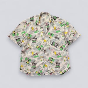Puede incluir: Camisa de manga corta con fondo beige claro y un estampado repetido de personajes de dibujos animados, palmeras y formas geométricas en verde, negro y amarillo. La camisa tiene cuello clásico y un bolsillo en el pecho.