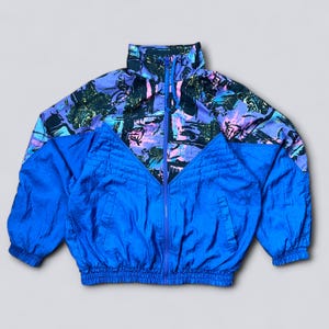 Op de afbeelding: Een vintage blauw en patroon trainingsjack met een volledige ritssluiting. De jas heeft een levendig ontwerp met blauwe, paarse en zwarte kleuren. De jas heeft een hoge kraag en geribde manchetten en zoom.