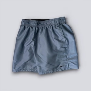 Puede incluir: Unos pantalones cortos deportivos de color gris claro con un acabado de tela suave y ligeramente reflectante, una cintura elástica y un pequeño logotipo Nike swoosh negro bordado en la pierna derecha. Tienen una abertura lateral.
