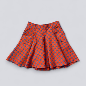 May include: A flared skirt in a vibrant orange fabric with a repeating blue geometric pattern. The skirt features two front pockets and a button closure. The skirt is a children's clothing item.